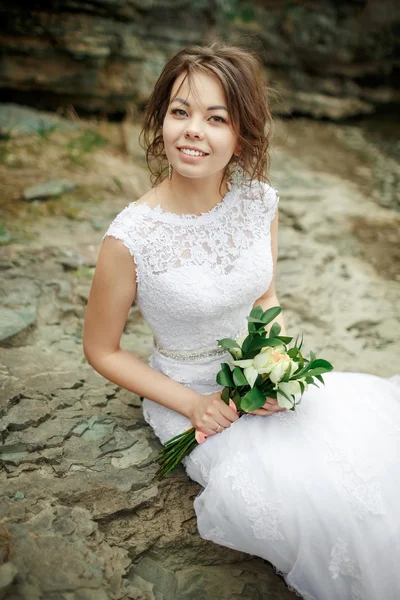 Bela noiva com cabelo desgrenhado em pedras entre pedras, buquê de casamento em mãos. Retrato sincero . — Fotografia de Stock