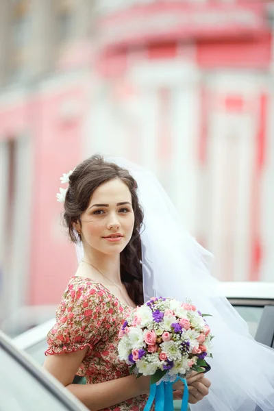 The bride in simple retro dress with floral pattern, already wearing veil, holding wedding bouquet, purple flowers, poses out of car, vertical frame. Looking straight at camera. — Stock Fotó