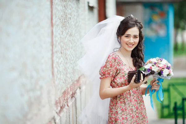 A noiva em um vestido retro simples com padrão floral, já vestindo véu, buquê de casamento e bolsa, posando fora de casa, olhando diretamente para a câmera, sorrindo . — Fotografia de Stock