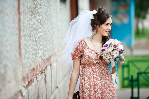 The bride in simple retro dress with floral pattern, already wearing veil, wedding bouquet and handbag, posing outside house, looking to side.