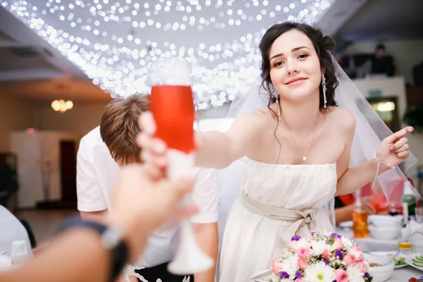 Beautiful young bride in white dress playfully raised glass of champagne and want to make a toast, clink glasses. Wedding banquet at restaurant, ceiling garlands. Celebrating. Cheers. — 图库照片