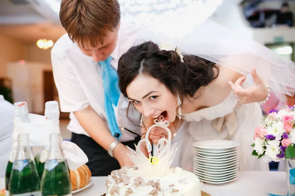 Impatient beautiful bride in white dress leaned forward quickly and wants to try the wedding cake, groom wearing turquoise tie stands nearby. Celebration, cake cutting. Wedding banquet at  restaurant. — 图库照片