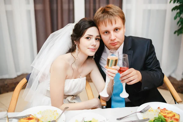 Newlyweds at the wedding table sitting together, posing with wine. Attractive beautiful bride, kind eyes, groom wearing suit and turquoise tie. Celebration, reception in restaurant is over. — 图库照片