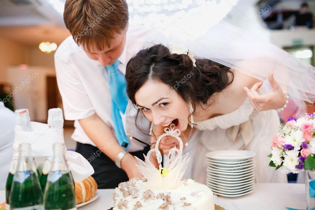 Impatient beautiful bride in white dress leaned forward quickly and wants to try the wedding cake, groom wearing turquoise tie stands nearby. Celebration, cake cutting. Wedding banquet at  restaurant.