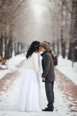 Asian bride and groom kissing in the middle of snowy winter alley. Young man wearing gray coat, fur hat, lady dressed with white wedding gown. Cold season warm dress.