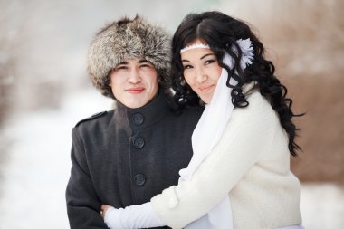 Beautiful wedding couple, asian bride and groom embraced. Young man in winter coat, fur hat, lady wearing dress with sheepskin, veil. Cold season warm clothing. Close-up portrait.