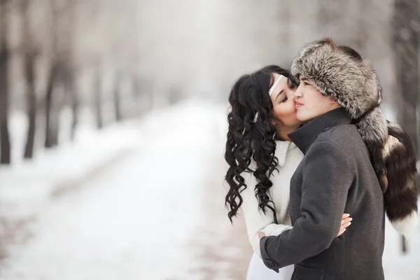 Exotic asian bride and groom kissing in middle of snowy winter alley. Young man wearing coat, fur hat, woman dressed by beautiful wedding gown with sheepskin. Cold season warm clothing. Copy space for — Stock Photo, Image