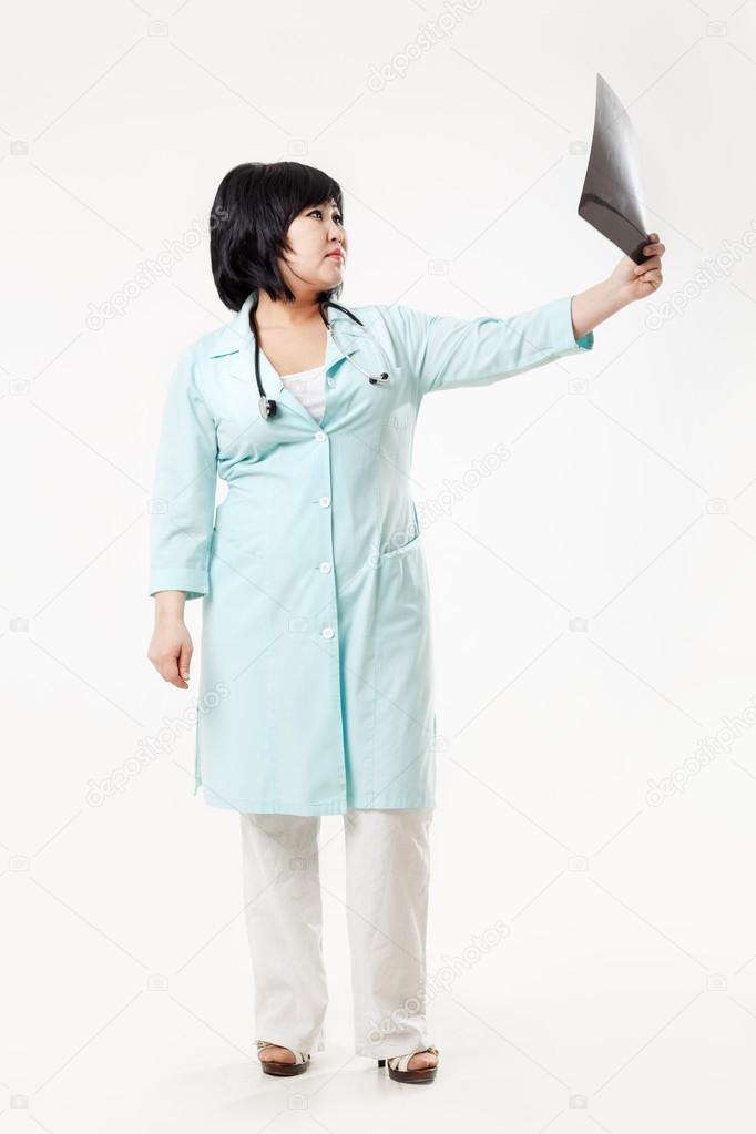 Curvy woman doctor standing considers the results of radiology department, x-ray skull picture outstretched arm, in turquoise medical gown with stethoscope. Asian appearance, dark short hair.