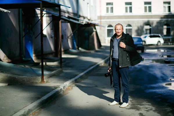 Brutaler Straßenfotograf mit der Kamera in der Hand, der durch die Straßen der Stadt flaniert. Porträt in voller Höhe. im Hof Mehrfamilienhäuser, Fotorucksack. — Stockfoto