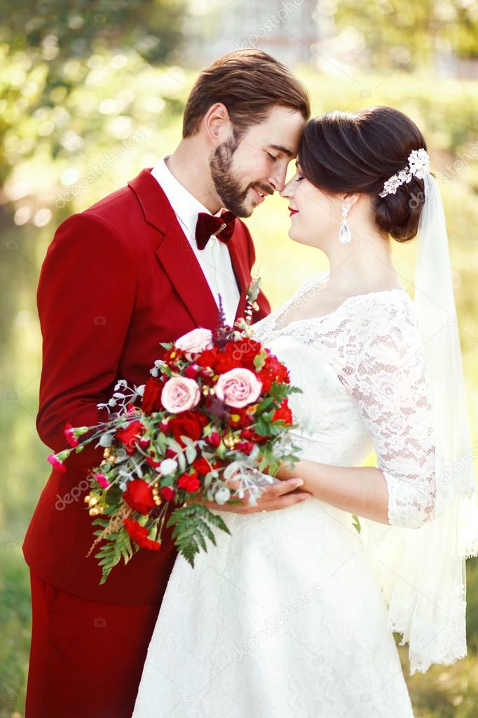 Bride and groom embracing, wedding couple, dark red color marsala style design. Suit with maroon bow tie, white dress, bridal bouquet. Professional makeup. Eternal love, tenderness, beauty concept.