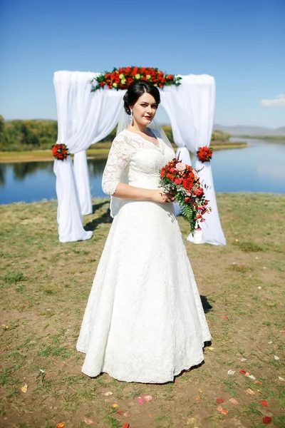 Noiva durante a cerimônia ao ar livre com buquê no rio, arco de rosas e pano branco . — Fotografia de Stock