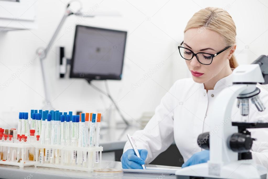 Professional young scientist is recording her research on the paper. She is sitting at the desk and writing with concentration in lab
