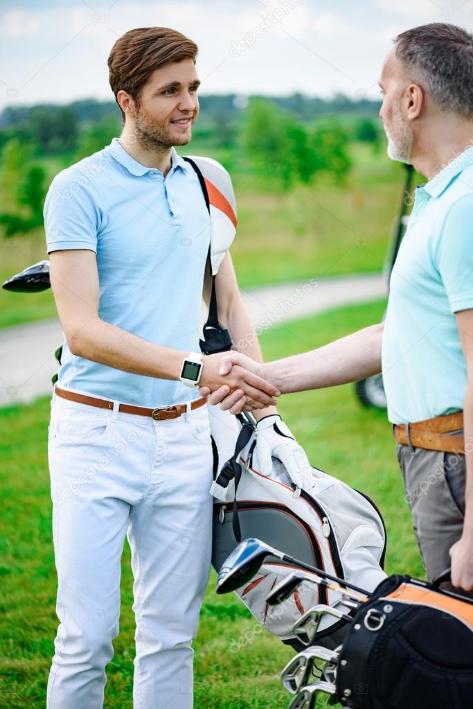 Thanking for game. Smiling young golfers shaking hands with his partner after playing together, holding golf backs