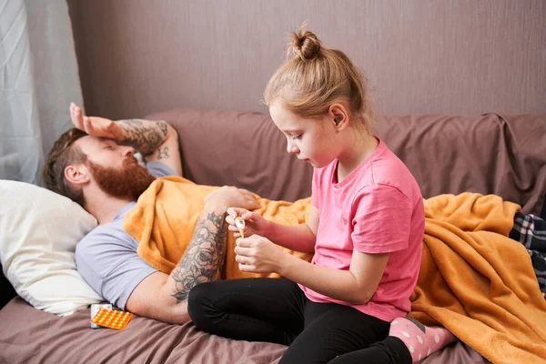 Hombre cubierto de manta está acostado en el sofá mientras su hija está tomando su temperatura — Foto de Stock