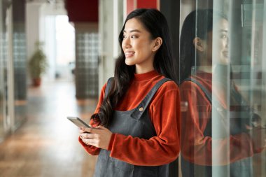 Gülümseyen, siyah saçlı, şık kıyafetler giyen, elinde akıllı bir telefon tutan bir kadın.