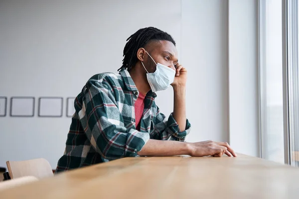 Sad young man in a medical mask looking through the window