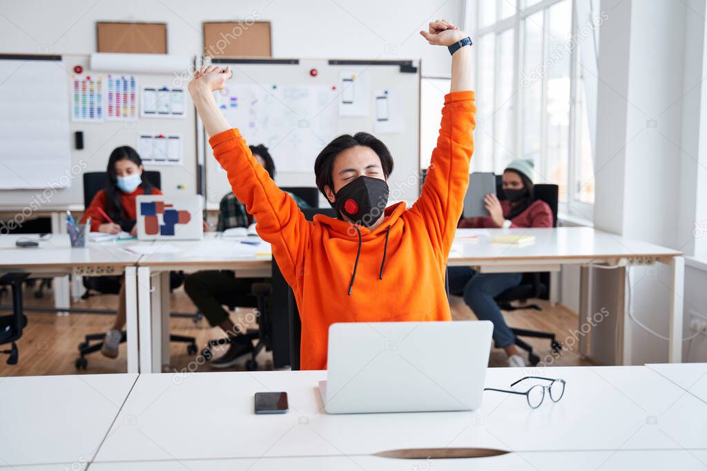 Student man wearing protective mask stretching while working with laptop computer at classroom