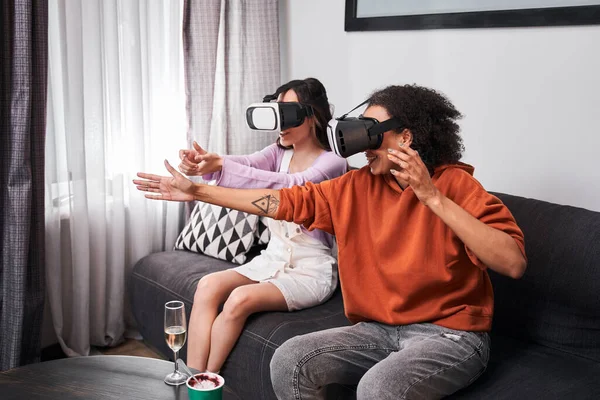 Las mejores amigas sentadas en un sofá acogedor y usando gafas de realidad virtual — Foto de Stock