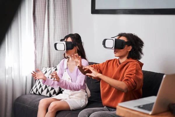 Las mejores amigas sentadas en un sofá acogedor y usando gafas de realidad virtual — Foto de Stock