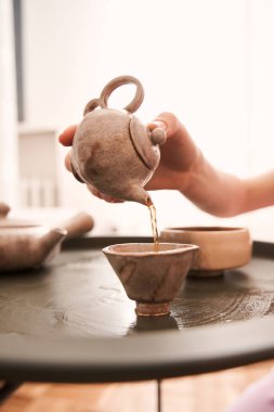 Yerde oturan bir kadın, elinde çaydanlıkla fincanlara çay dolduruyor.