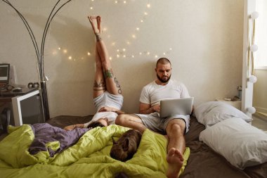 Kadın, dizüstü bilgisayarla çalışırken kocasının yanında bacaklarını kaldırıyor.