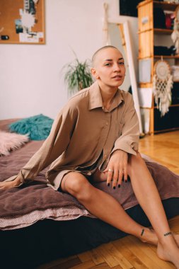 Kısa saçlı dövmeli kız şiltede oturuyor ve kameraya poz veriyor.