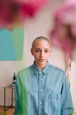 Kısa saçlı ve piercingli bir kadın çiçeklerin arkasındaki dairesinde poz veriyor.