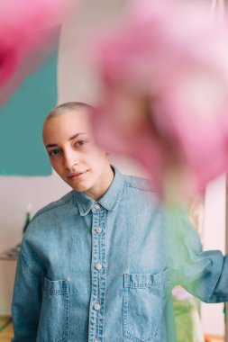 Kısa saçlı ve piercingli bir kadın çiçeklerin arkasındaki dairesinde poz veriyor.