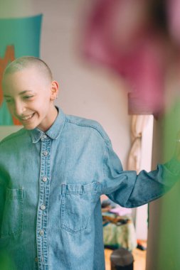 Kısa saçlı ve piercingli bir kadın şık bir kot gömlek giyiyor ve evinde poz veriyor.