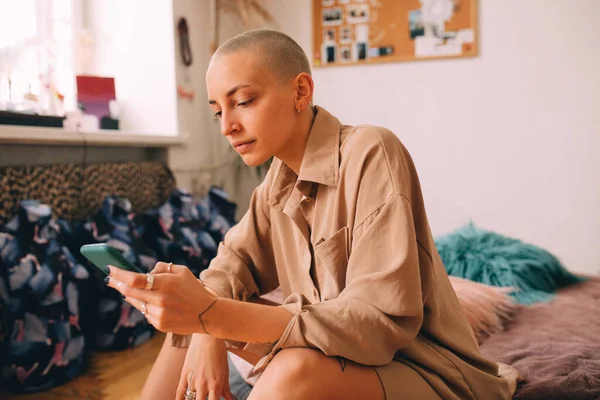 Menina com corte de cabelo curto sentado no colchão e olhando para o smartphone — Fotografia de Stock