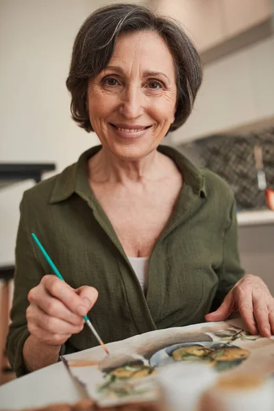 Woman smiling to the camera happily while sitting calmly at the table and drawing — Stock Photo, Image