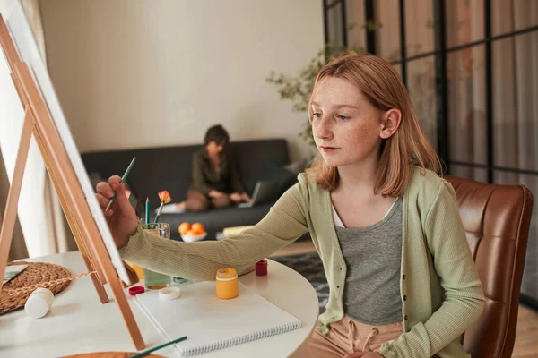 Girl artist sitting in front of easel in her kitchen and making picture — Stock fotografie