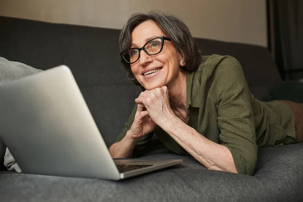 Жінка в окулярах лежить на дивані зі своїм ноутбуком і мріє про щось — стокове фото