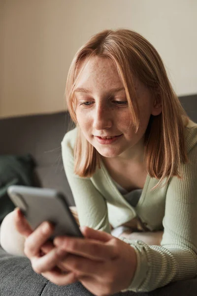Chica pecosa mirando la pantalla del teléfono inteligente mientras se sienta en el sofá — Foto de Stock