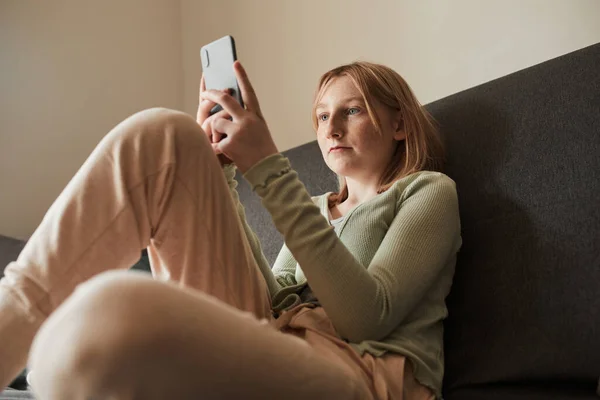 Chica de la escuela sentada en el sofá y el uso de su teléfono móvil mientras navega por Internet — Foto de Stock