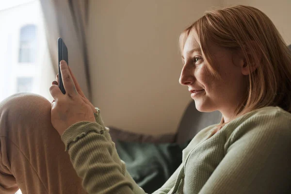 School girl sitting at the couch and using her mobile phone while browsing internet — Stock fotografie