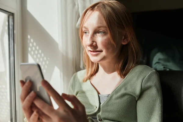 Ginger girl looking at the window while sitting at the windowsill with her smartphone — Stock fotografie