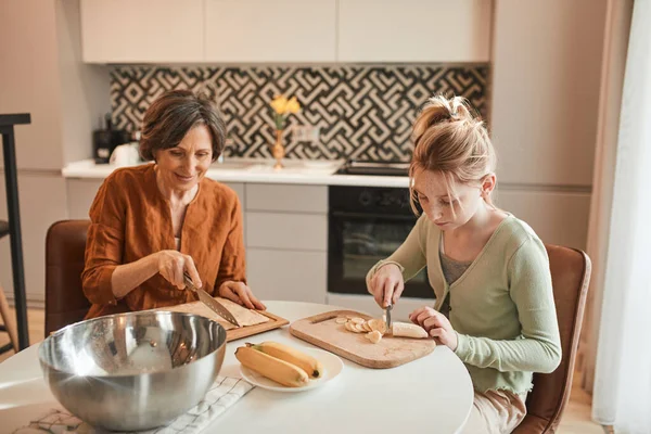 Chica picando con el cuchillo plátano fresco mientras hornear rosquillas con su abuela —  Fotos de Stock