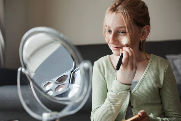 Girl looking at the mirror and preparing makeup with a powder and brush while playing — Stockfoto