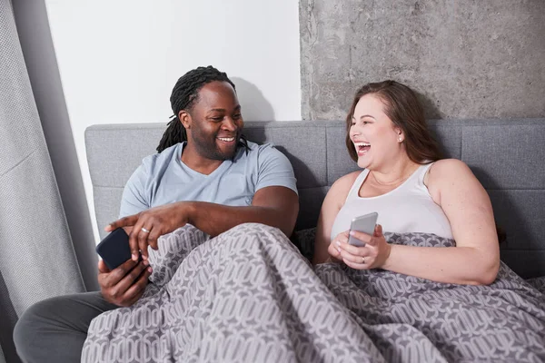 Nő ül az ágyban a férjével, és néz valami vicces okostelefonok — Stock Fotó