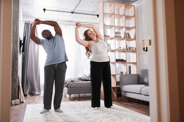 Homem multirracial e sua namorada caucasiana em roupas domésticas fazendo exercícios matinais — Fotografia de Stock