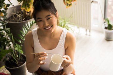 Evde resim yaparken elinde fincan ve boya fırçası tutan kadın sanatçı.