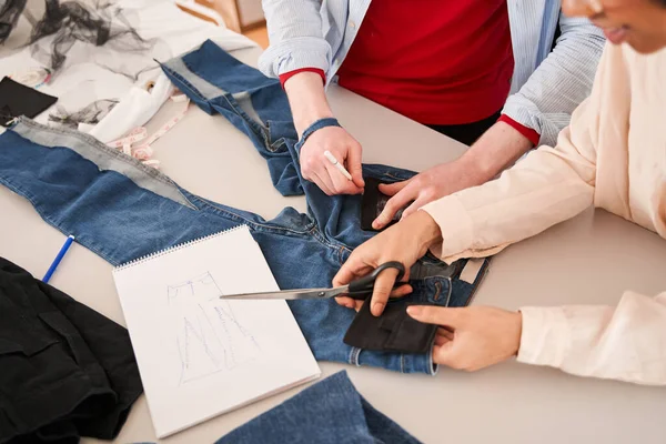 Colegas examinando jeans e desenhando esboços enquanto re costura roupas velhas — Fotografia de Stock