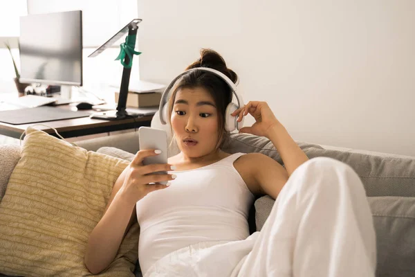 Surprised young woman chilling on couch while enjoying podcast at home