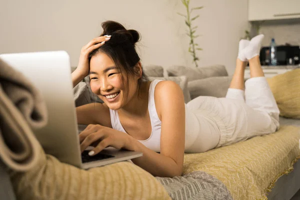アジアのブルネットの女性は家のベッドの上に寝そべっている間にラップトップで働く — ストック写真