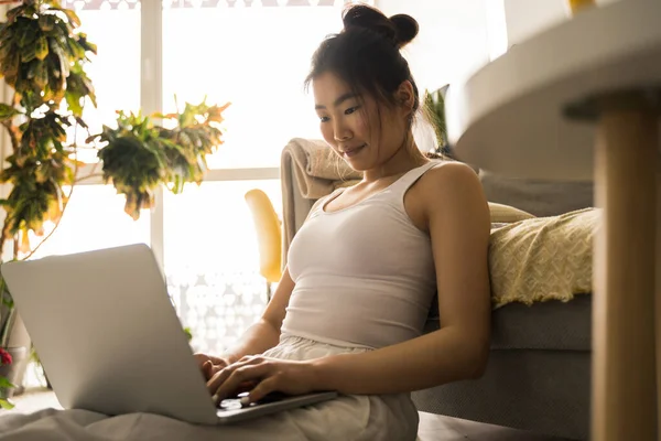 Mujer asiática sentada en el suelo mientras escribe correo electrónico en el portátil —  Fotos de Stock