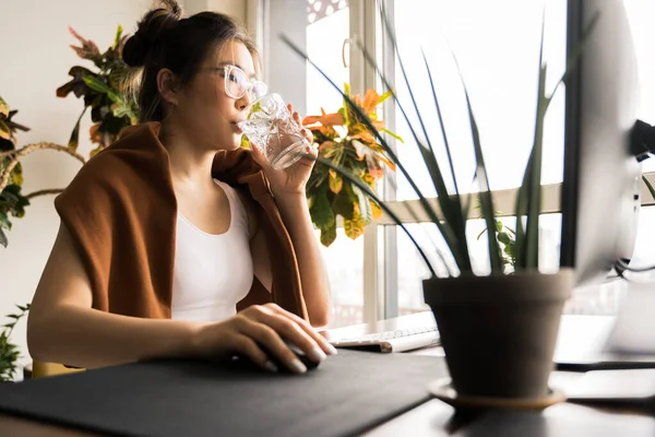 Жінка п'є воду, сидячи за столом зі стаціонарним комп'ютером — стокове фото