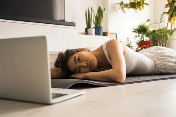 Mujer tendida en la alfombra en el suelo y viendo clases de yoga en el portátil —  Fotos de Stock