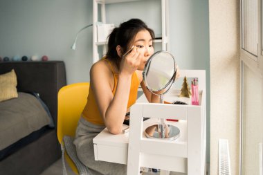 Uzun saçlı bir kadın masada otururken günlük doğal makyaj yapıyor.