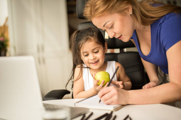 Mom standing near the desk and helping with studying to her little daughter